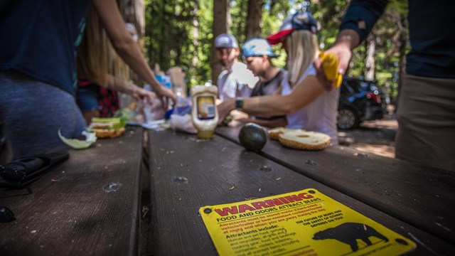 A group sits in a campground preparing a meal, in the foreground a bear safety sign.