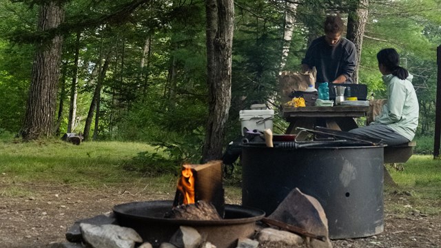 Two campers eat at a wooden picnic table in a campsite along the East Branch Penobscot River.