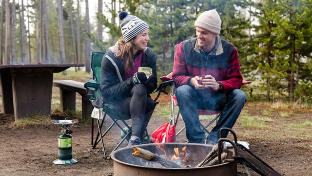 A man and a woman sit by a campfire in a campsite and drink coffee.